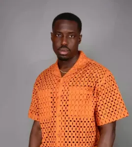 A man wearing an orange, short-sleeved, perforated shirt stands against a plain backdrop. British-Ghanaian star Mark Asari scores big with ‘Energy’ in new Netflix movie “Beverly Hills Cop: Axel F.”