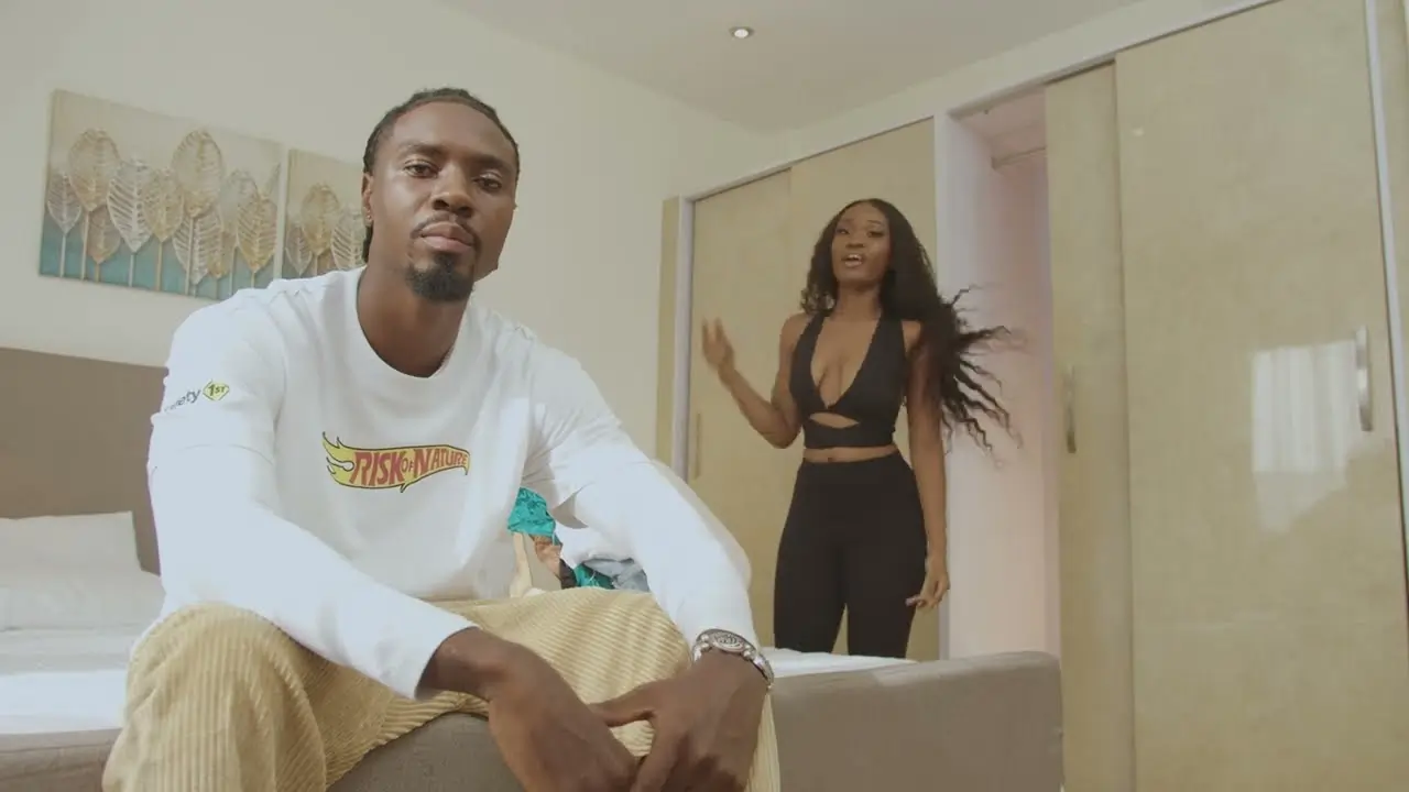 A man sitting on the edge of a bed looking at the camera while a woman enters the room in the background. Thumbnail for "Beeztrap KOTM - Fly Girl feat. Oseikrom Sikanii (Official Video)"